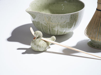 Green ceramic bowl with a swan-shaped spoon holder and wooden sticks on a white surface.